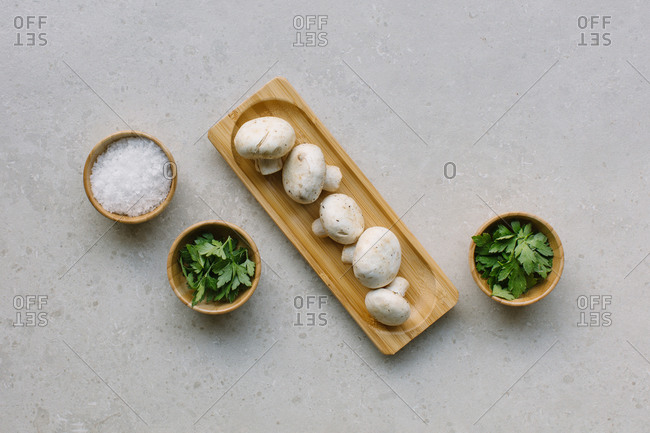 Top view of fresh champignons served on wooden plate on table near green parsley and salt in bowls