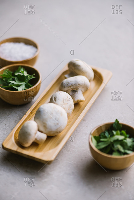 Fresh mushrooms served on wooden plate on table near green parsley and salt in bowls