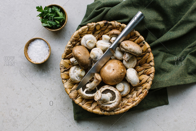Top view of fresh champignons in wicker bowl placed on table in a green napkin with salt and parsley in kitchen