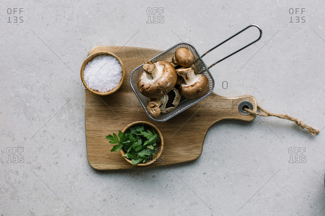 From above of raw mushrooms in metal container on table near bowls with salt and parsley placed on wooden cutting board