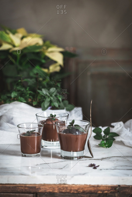 Still-life shoot of chocolate and mint mousse served inside glasses, placed on marble table against decorated background