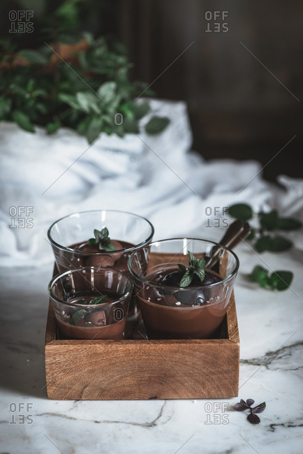 Still-life shoot of chocolate and mint mousse served inside glasses, placed on marble table against decorated background