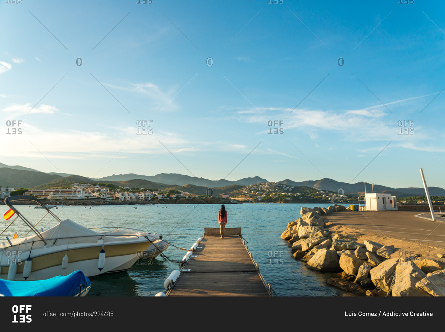 Back view of unrecognizable female traveler standing on wooden pier near vessels and admiring picturesque view of small Llanca municipality with buildings and cloudy sky reflected in calm water in sunny day in Spain