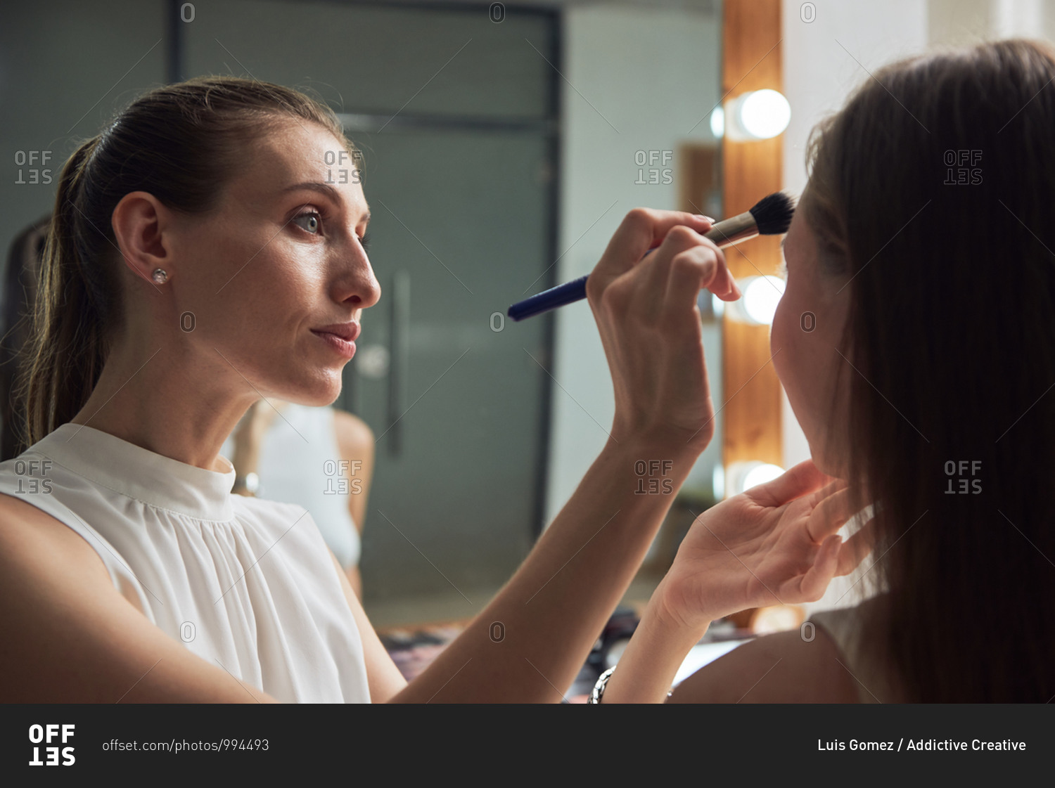Positive young female makeup artist applying powder on clients face by using brush against cozy big mirror with lights in contemporary beauty salon