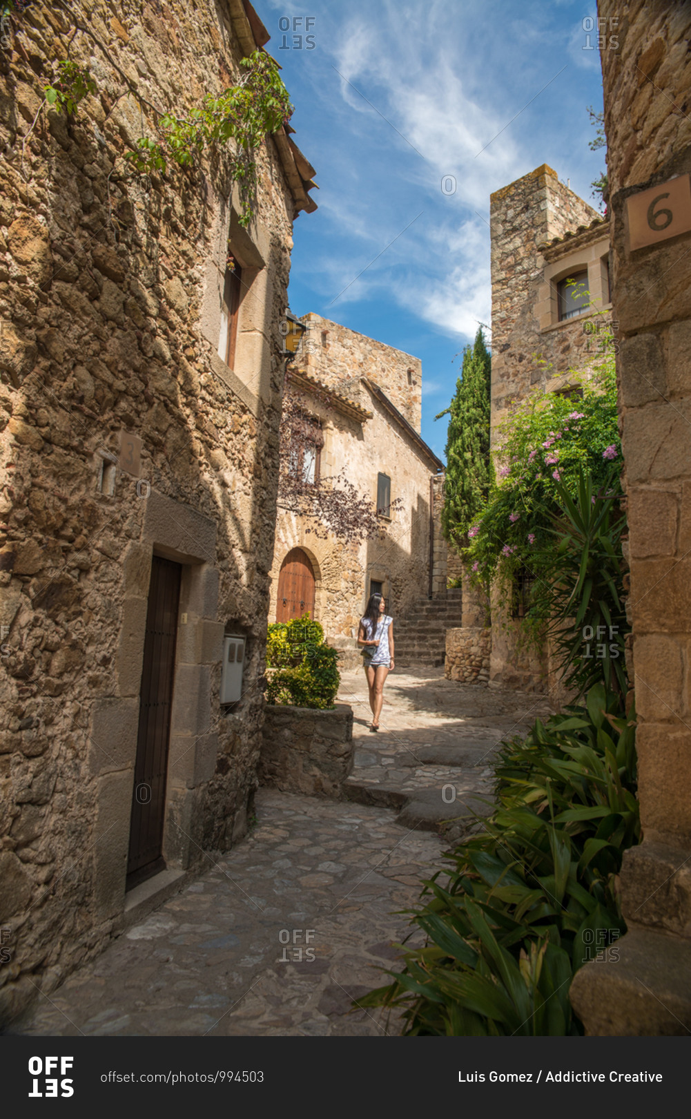 Young ethnic female tourist in casual clothes walking on narrow street among ancient stone buildings with green plants while visiting Tossa de Mar municipality in Catalonia in Spain