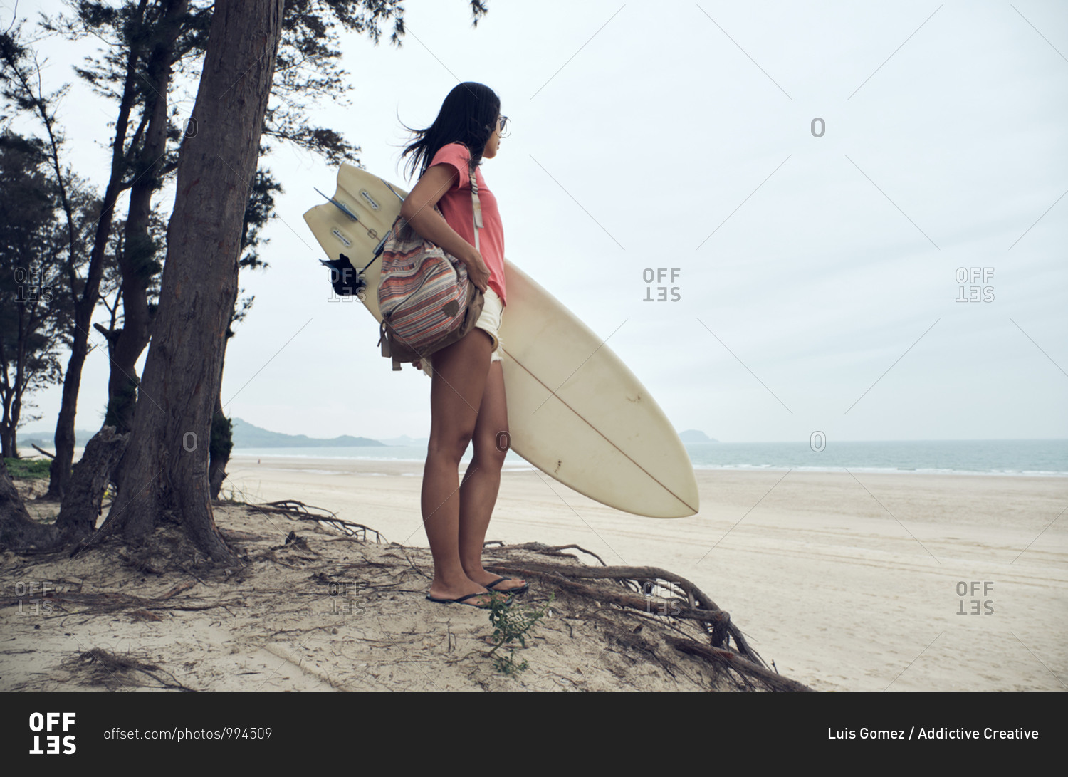 Side view of young Asian female surfer in summer outfit walking on sandy beach and carrying surfboard against calm blue sea looking away