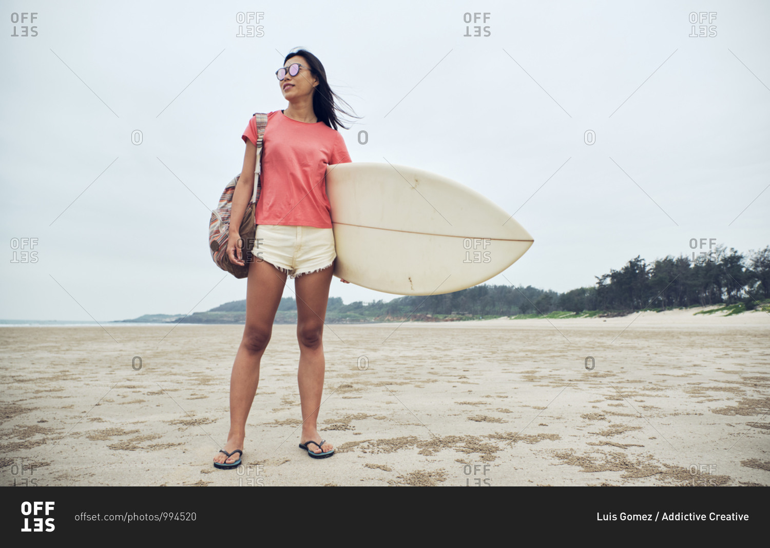 Full length young Asian female surfer in summer outfit walking on sandy beach and carrying surfboard against calm blue sea looking away