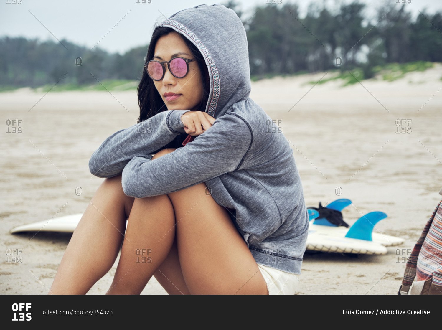 Crop contemplate Asian female in hoodie and sunglasses sitting on sandy beach near surfboard and looking away thoughtfully