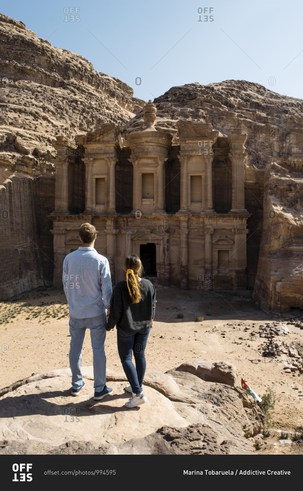 Back view of traveling couple holding hands standing on rocky hill while admiring amazing scenery of aged monastery in Jordan