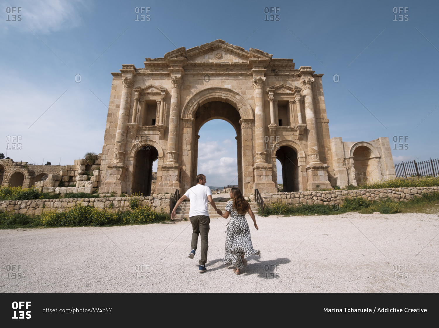 Back view of traveling girlfriend and boyfriend holding hands and cheerfully running to historic arch in Jerash