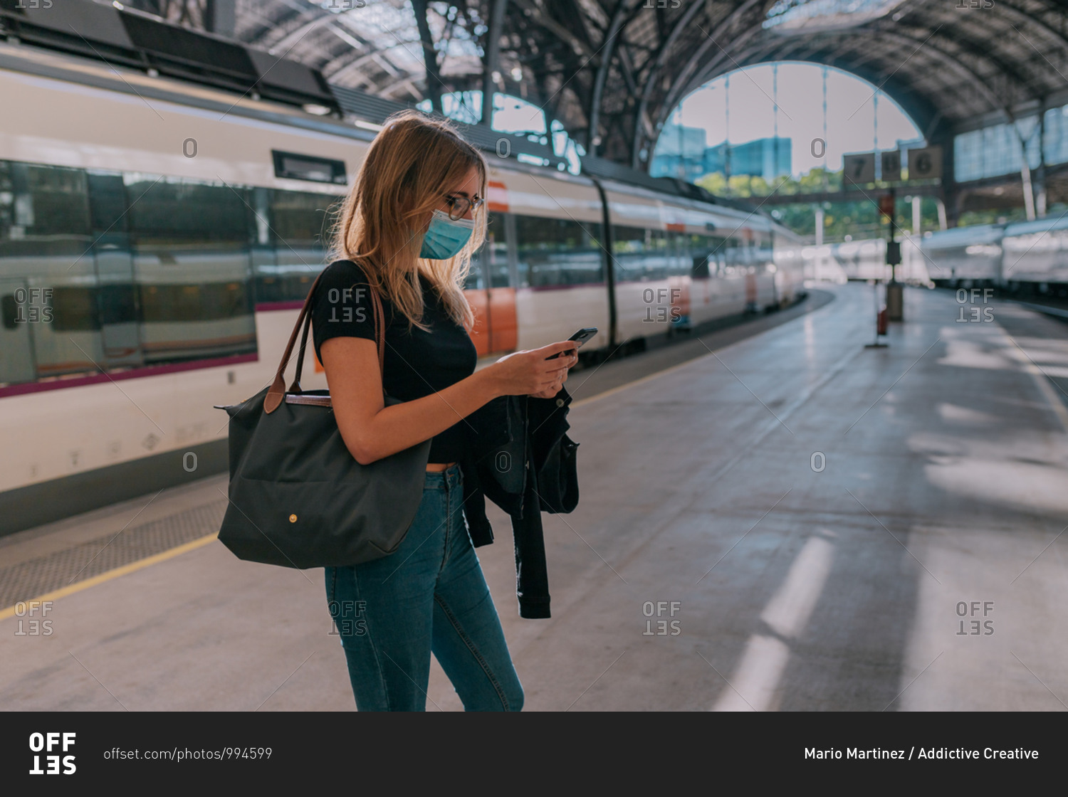 Young woman wearing a face mask waiting at the train station and looking at the smartphone during a pandemic