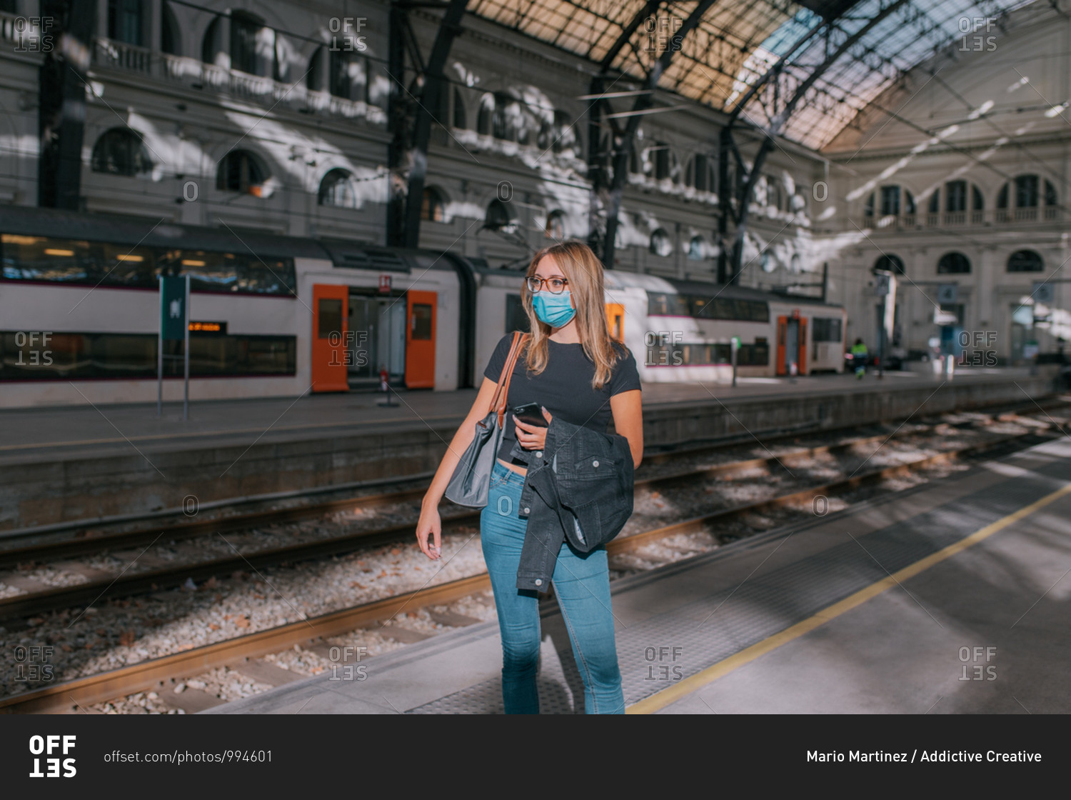 Young woman wearing a face mask waiting at the train station with a smartphone during a pandemic