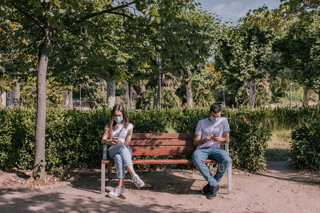 Couple at the park using the smartphone, wearing face masks and respecting social distancing during a pandemic