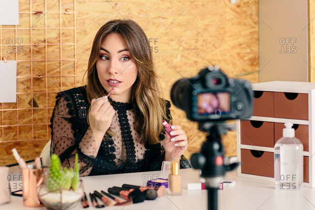 Charming female influencer in elegant wear applying lipstick while looking at camera making a tutorial recording video on professional camera
