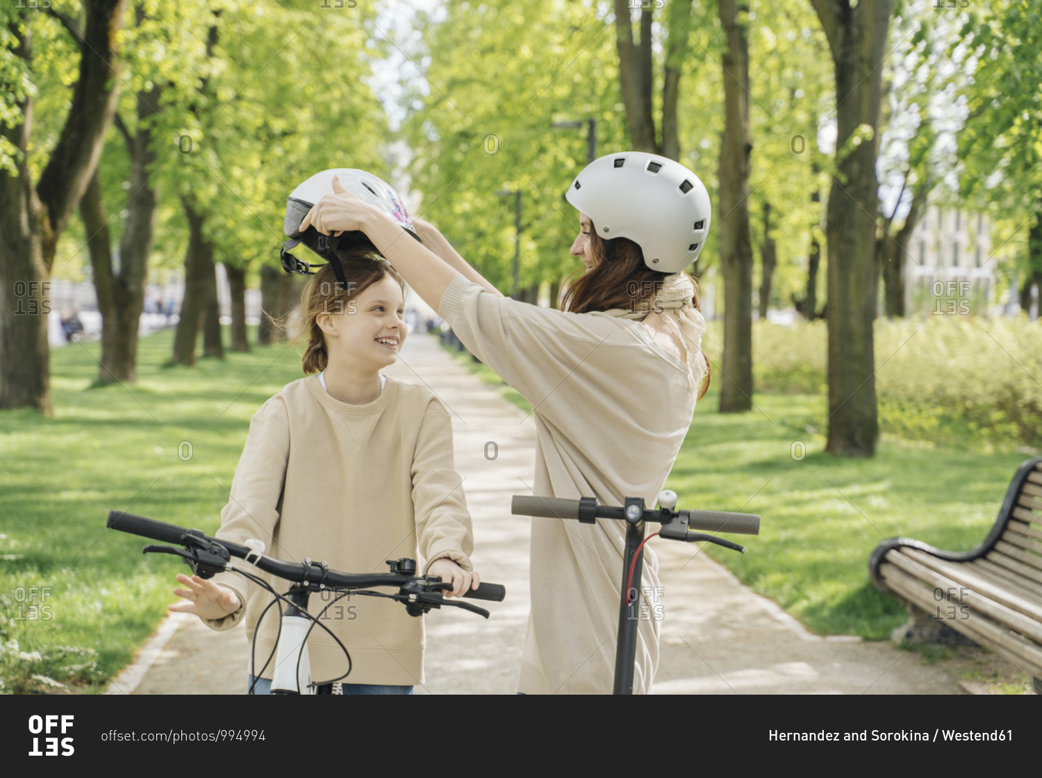 Mother wearing helmet to her daughter while standing against trees in park