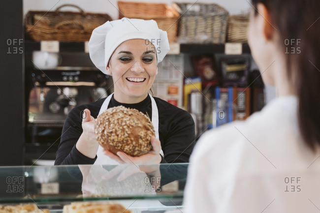 Happy female baker selling wholegrain bun to customer at bakery