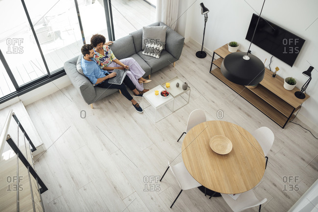 Multi-ethnic couple using laptop while sitting on sofa in living room of modern penthouse