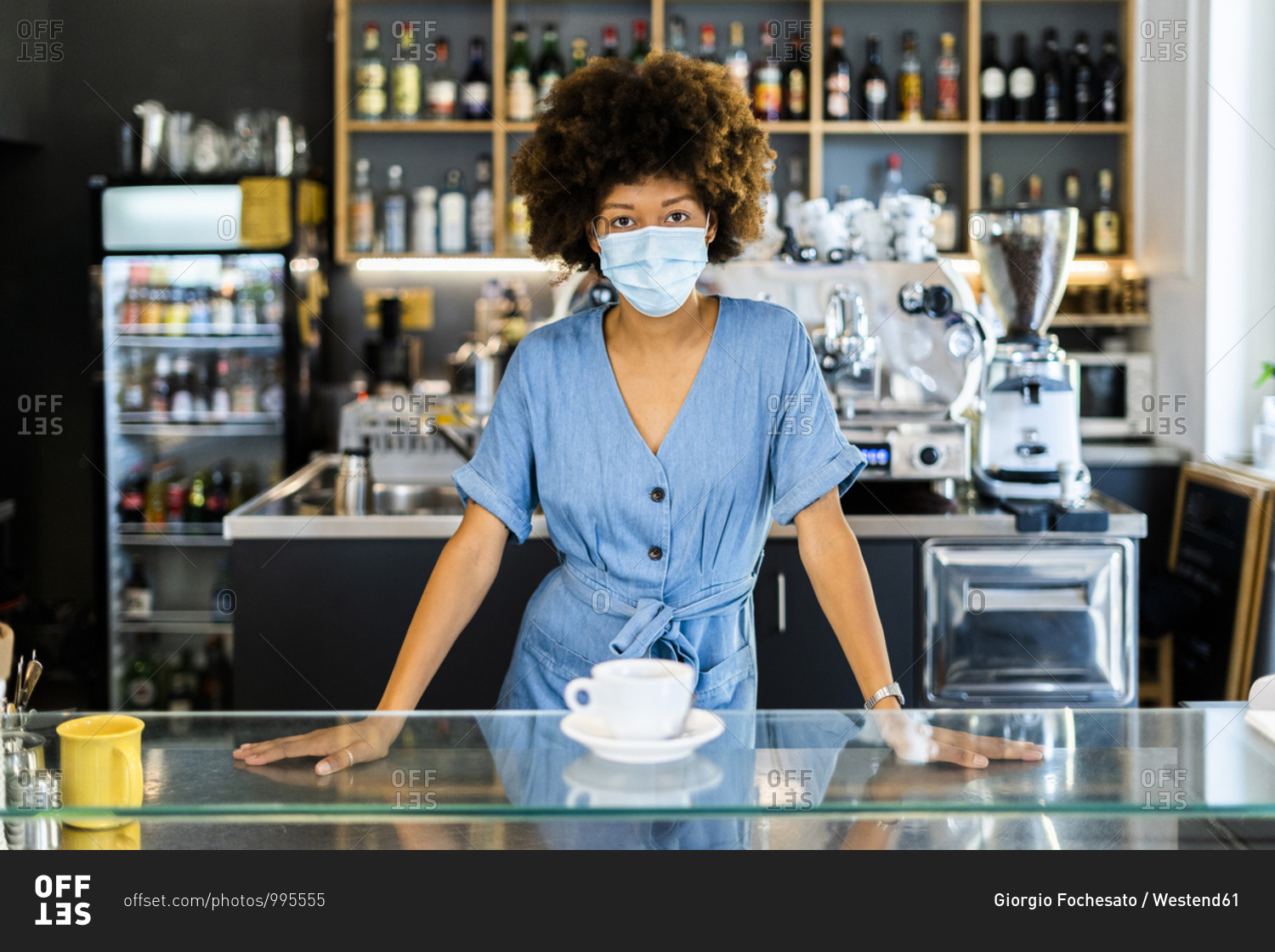 Young female owner wearing mask standing at cafe counter