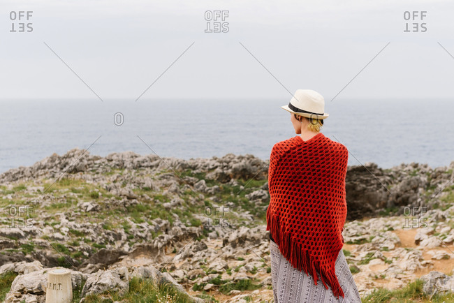 Back view of unrecognizable female traveler in stylish outfit and hat standing on edge of rocky cliff on seashore while exploring Asturian coast of Spain during summer vacation