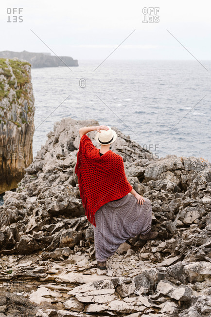 Full length back view of unrecognizable woman in stylish clothes standing on edge of rocky cliff against cloudy sky and admiring seascape while enjoying holidays on Asturian coast of Spain