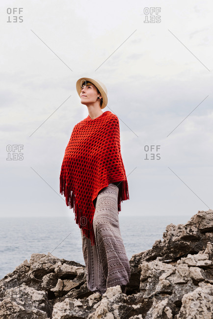 From below full length of woman in stylish clothes standing on edge of rocky cliff against cloudy sky and admiring seascape while enjoying holidays on Asturian coast of Spain