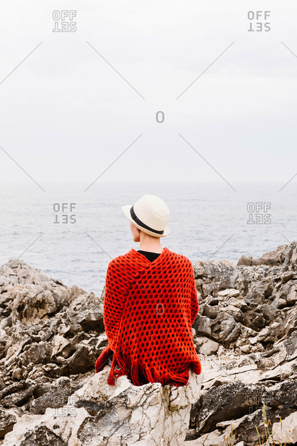Back view of unrecognizable woman in stylish clothes sitting on edge of rocky cliff against cloudy sky and admiring seascape while enjoying holidays on Asturian coast of Spain