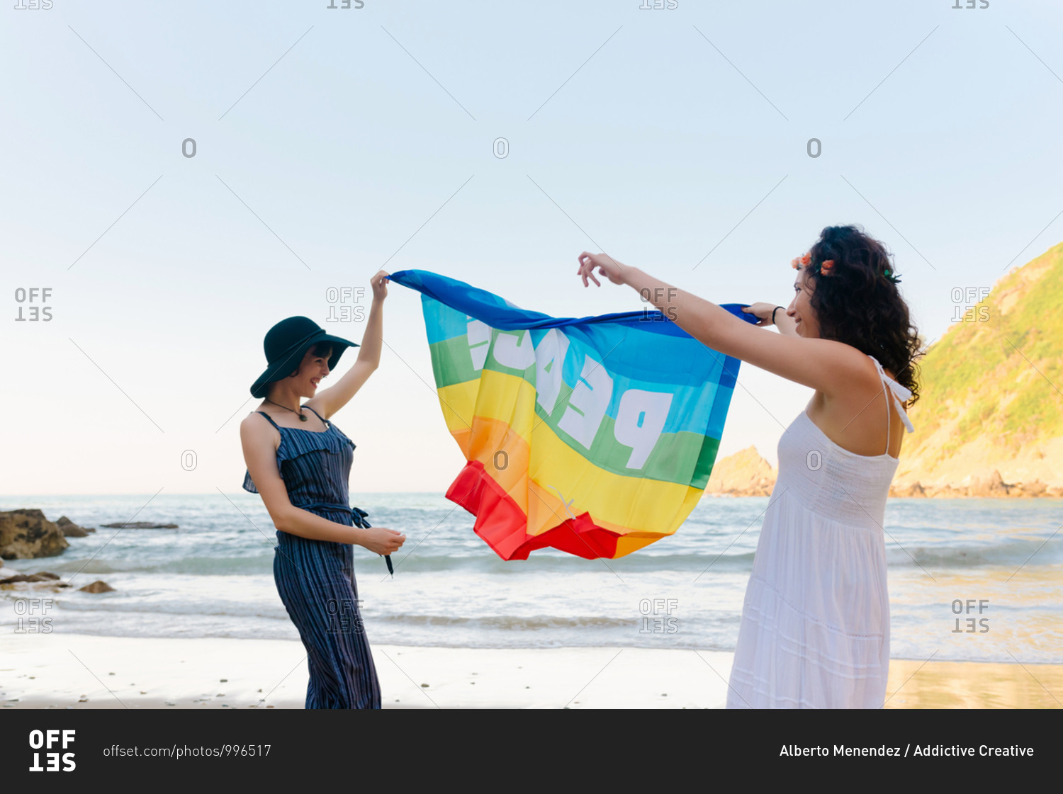 Side view of slim lady in hat and cheerful girlfriend in sundress raising colorful pride flag while standing on sandy shore near wavy ocean during vacation and looking at each other