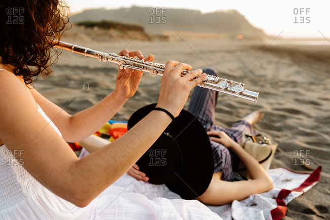Skilled female musician sitting on blanket on shore and playing flute for girlfriend while resting in evening