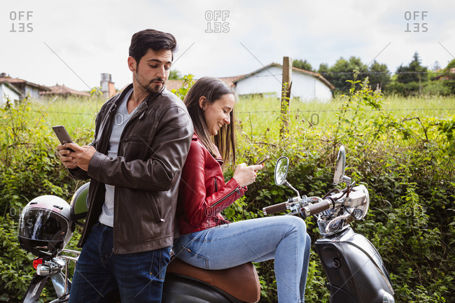 Side view of masculine guy using social media on cellphone while standing near content beloved sitting on motorcycle near lush green shrubs in rural zone under cloudy sky