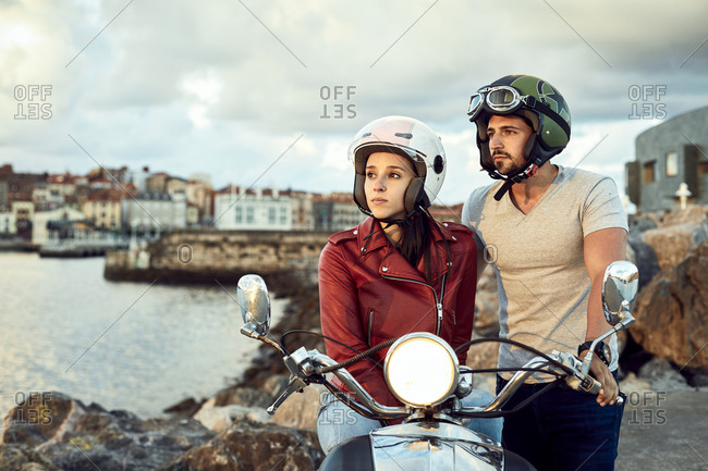 Thoughtful man in helmet standing near wistful girlfriend in leather jacket sitting on motorcycle with headlight on while contemplating picturesque river during vacation and looking away