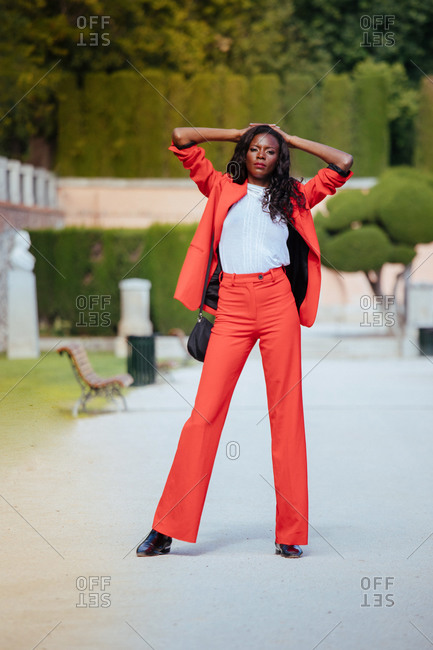 Slim African American woman in bright trousers and jacket standing on walkway leaned on hands near wooden bench and shrubs in city park and looking at camera