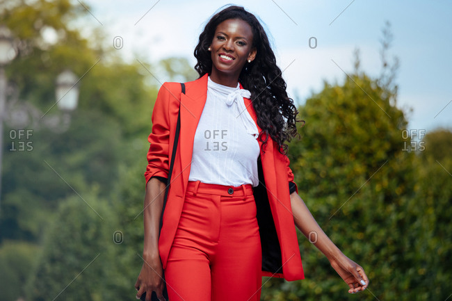Slim pensive African American female in colorful suit touching hair while looking at camera in city park near green shrubs in summer under cloudy sky