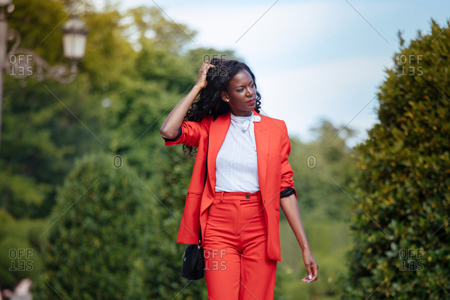 Slim pensive African American female in colorful suit touching hair while looking away in city park near green shrubs in summer under cloudy sky