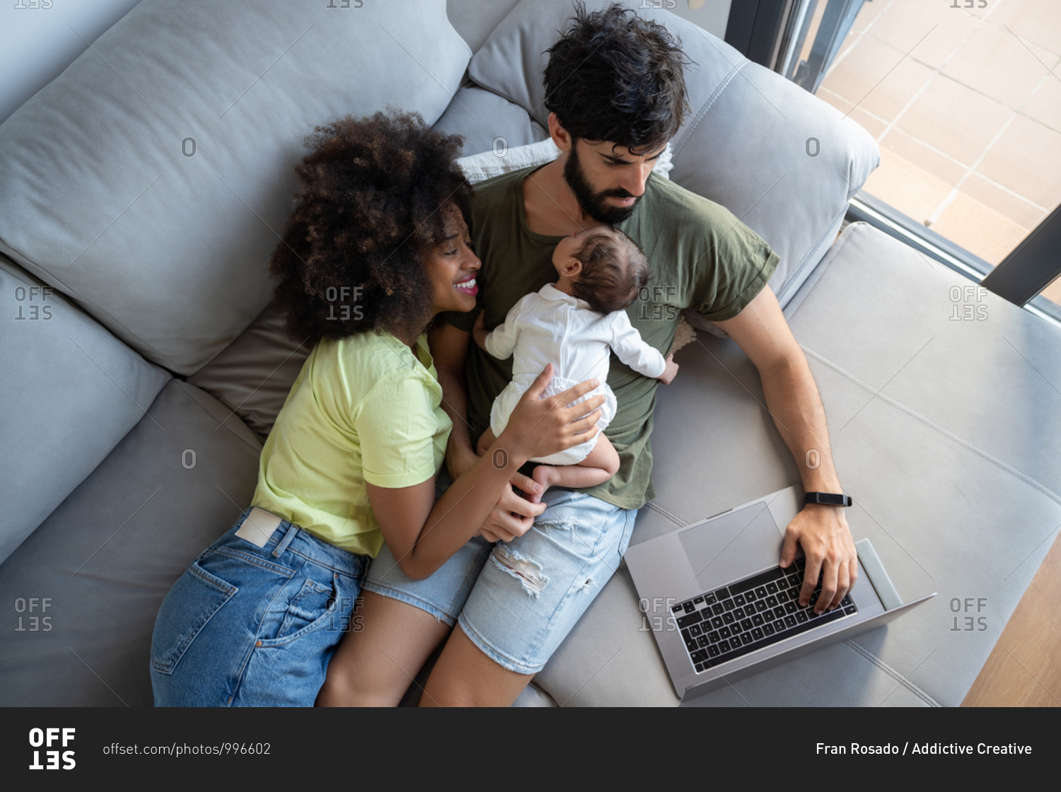 From above of black mother and cute infant lying on couch with Arab father working on laptop at home