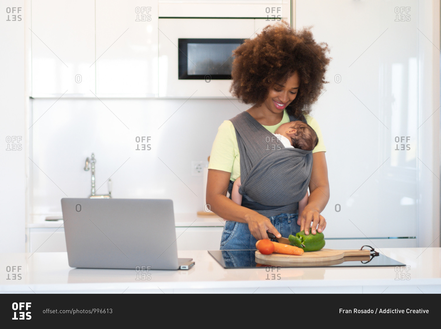 African American mother carrying sleeping infant in sling and cutting fresh vegetables in kitchen while preparing tasty lunch watching recipe on laptop