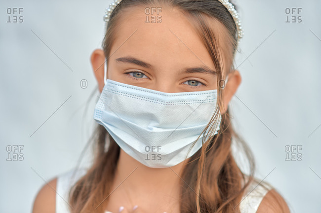 Preteen blond girl in a white dress wearing a protective mask because of the covid-19 pandemic, on the day of her first communion standing on white wall background