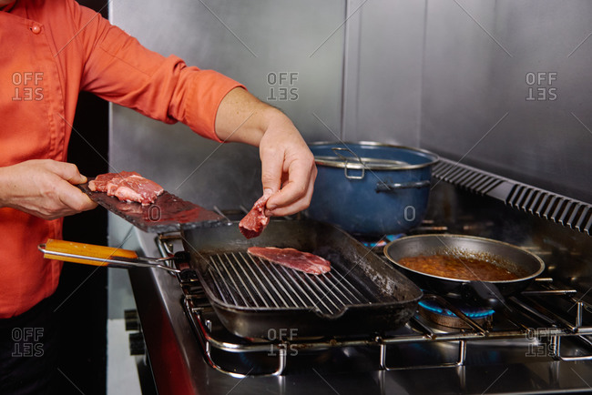 Crop unrecognizable chef frying thin beef slices on grill pan on stove while showing delicious surface of meat using tweezers in kitchen