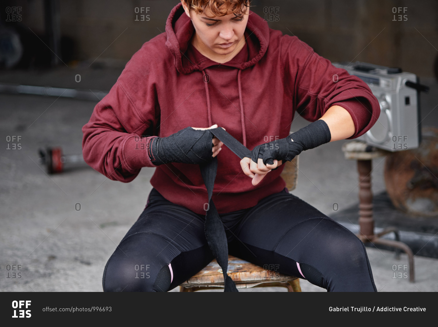 Crop concentrated female boxer in leggings and hoodie wrapping hands with black boxing wraps before boxing workout in shabby basement gym