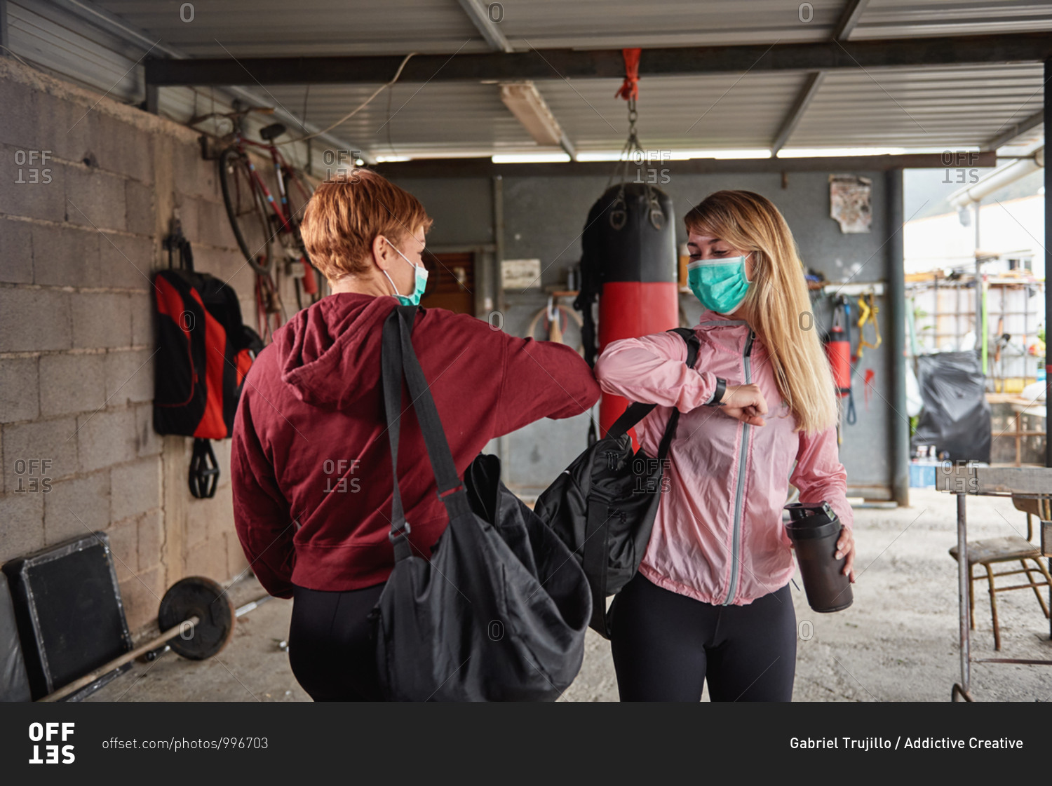 Sporty females in activewear with sports bags and protective face masks greeting each other with elbow bumps in rural garage gym during coronavirus outbreak
