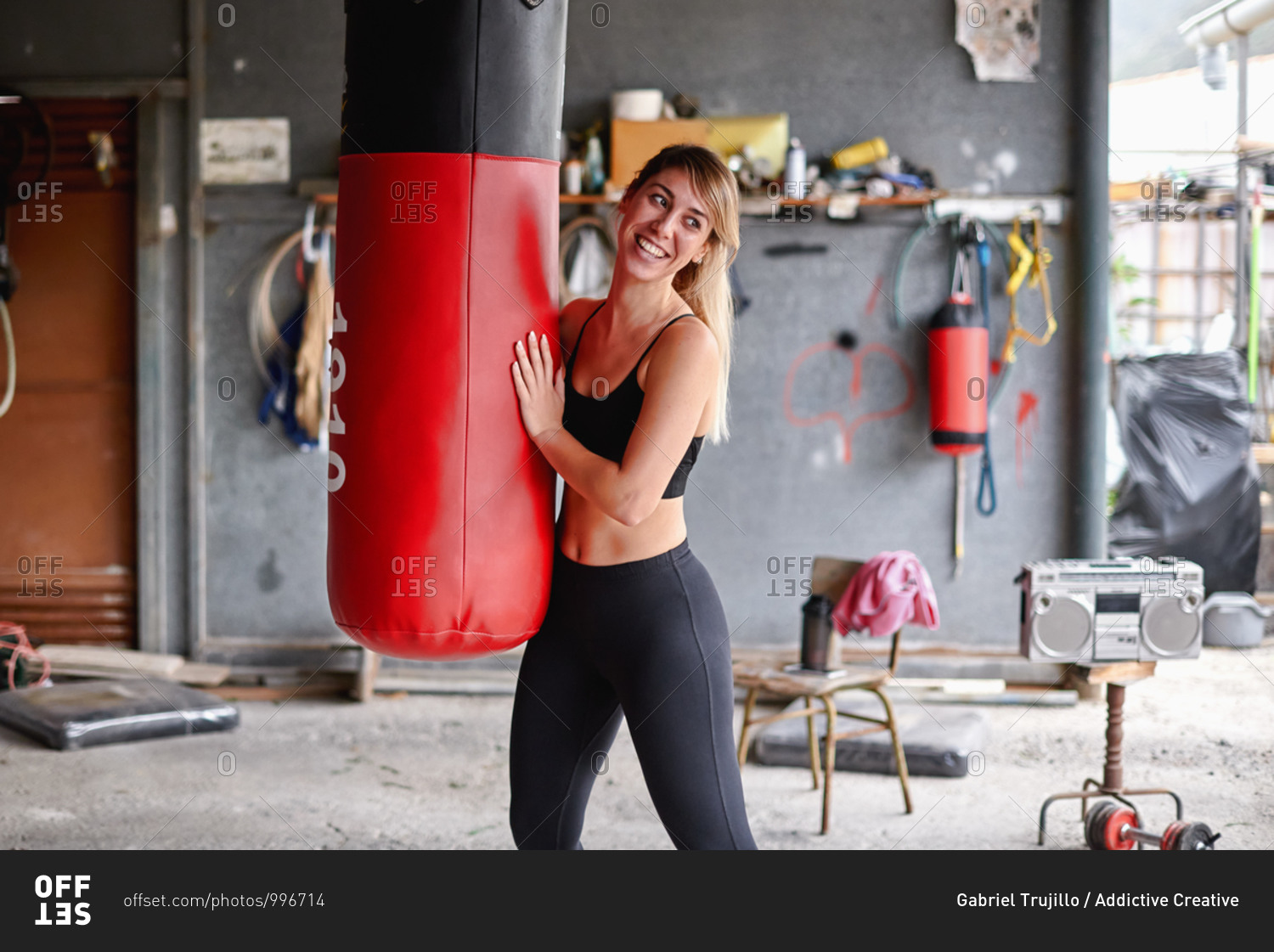 Positive young sportswoman wearing leggings and sports bra hugging punching bag and looking away happily after intense workout in messy rural garage gym