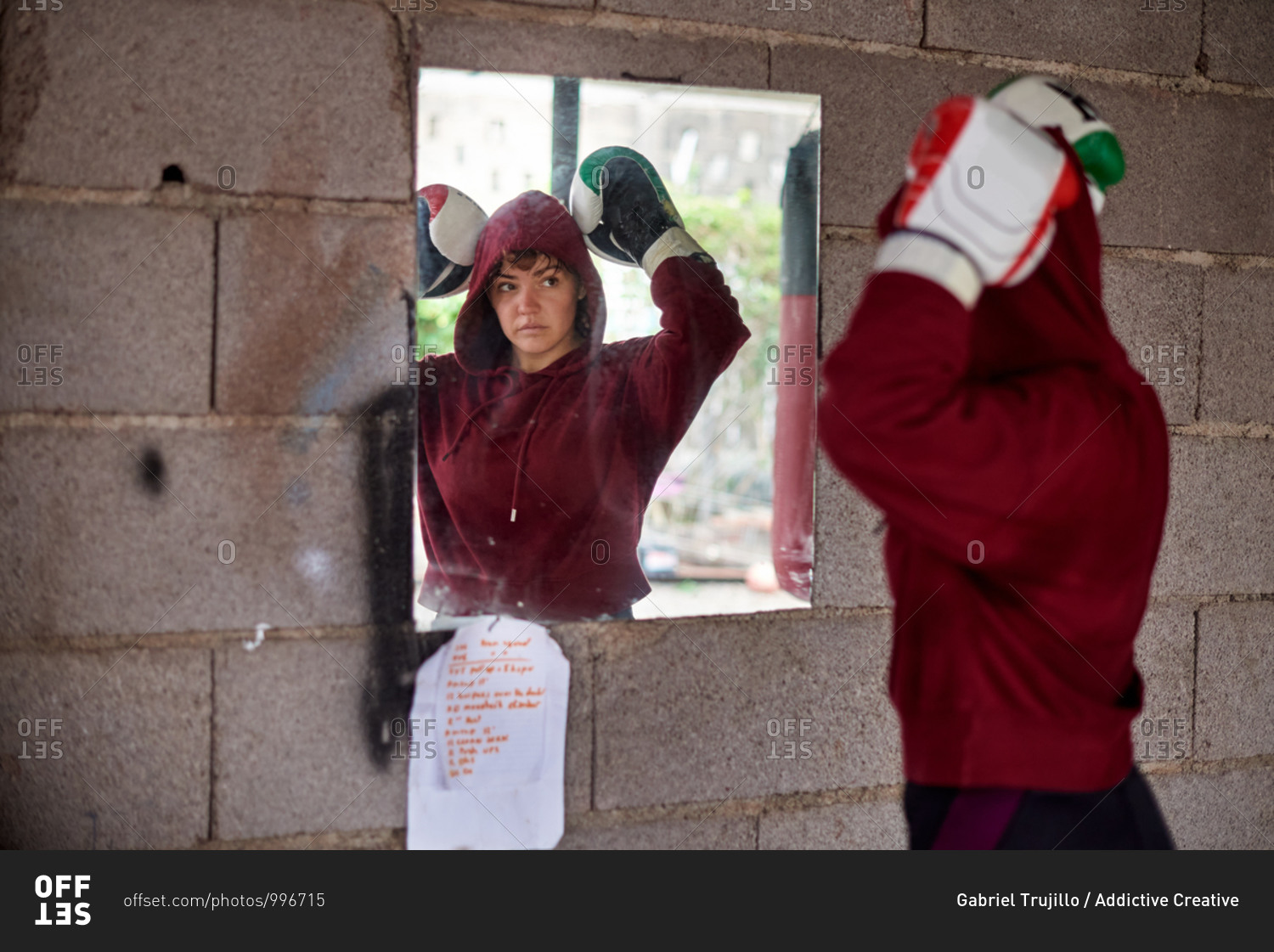 Serious sportswoman wearing hoodie and boxing gloves standing in double biceps pose and looking in mirror on rural shabby wall