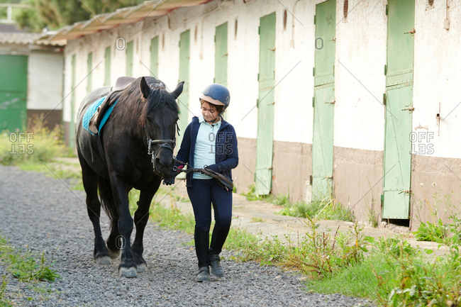Girl in rider clothes leading black stallion with saddle by reins while walking on pathway near stable in daylight and looking away
