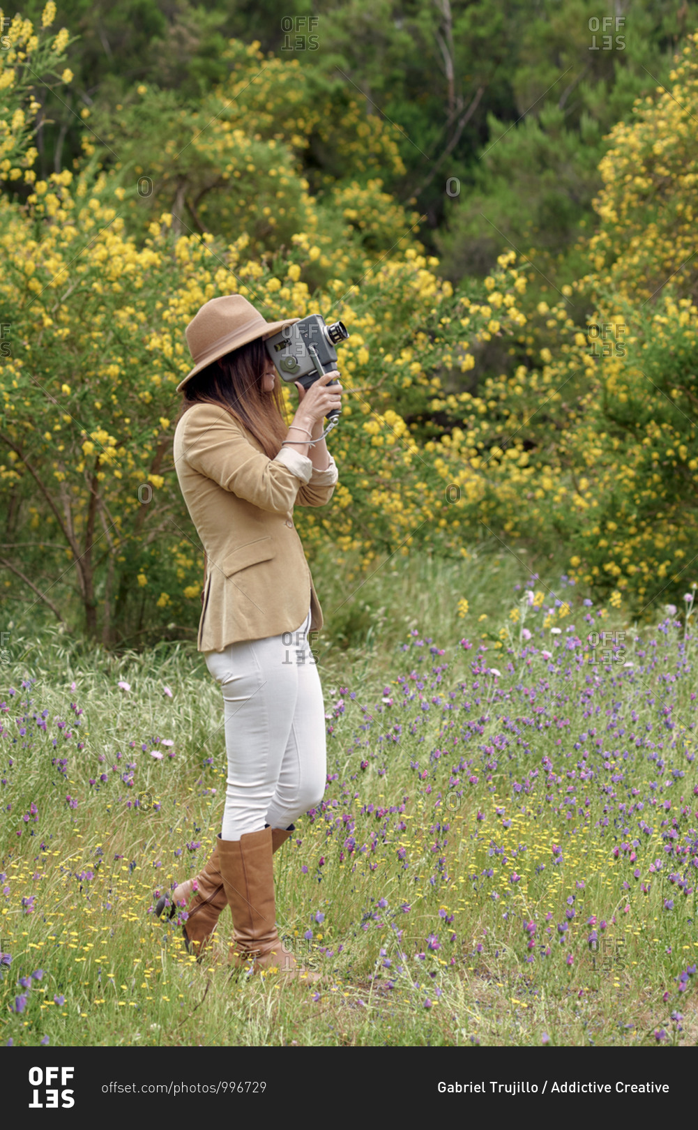 Female photographer in hat with closed eyes filming video with retro video camera while standing near trees with blooming yellow flowers in daylight
