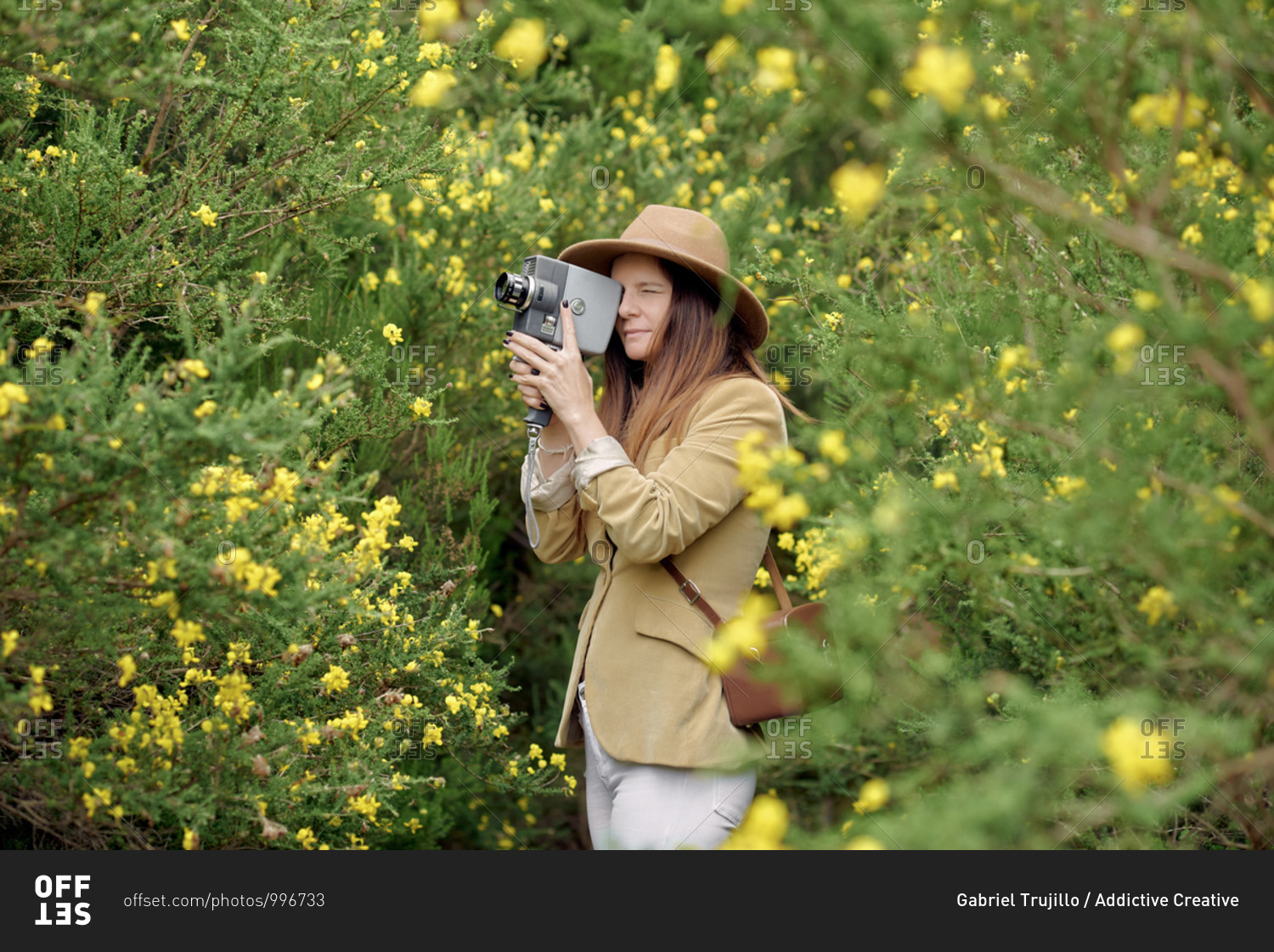 Smiling slim woman in casual wear and hat strolling with old video camera and leather case near bushes with colorful blooming flowers in countryside and looking away