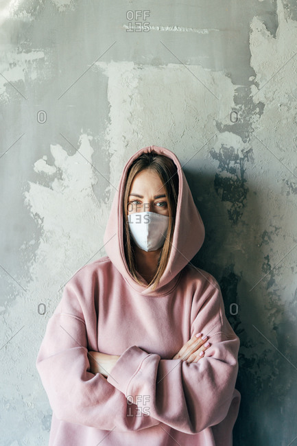 Unrecognizable discontent female in pullover and sterile mask standing with crossed arms near cement wall with shabby surface in flat and looking at camera during COVID 19 pandemic