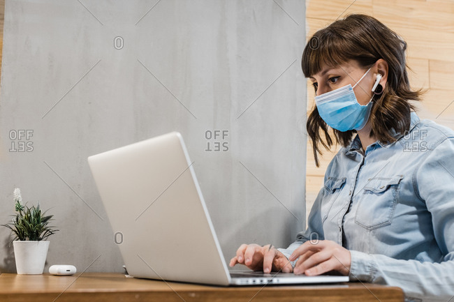 Focused female freelancer in denim shirt and medical mask using earbuds and working remotely in cafe using laptop during coronavirus epidemic