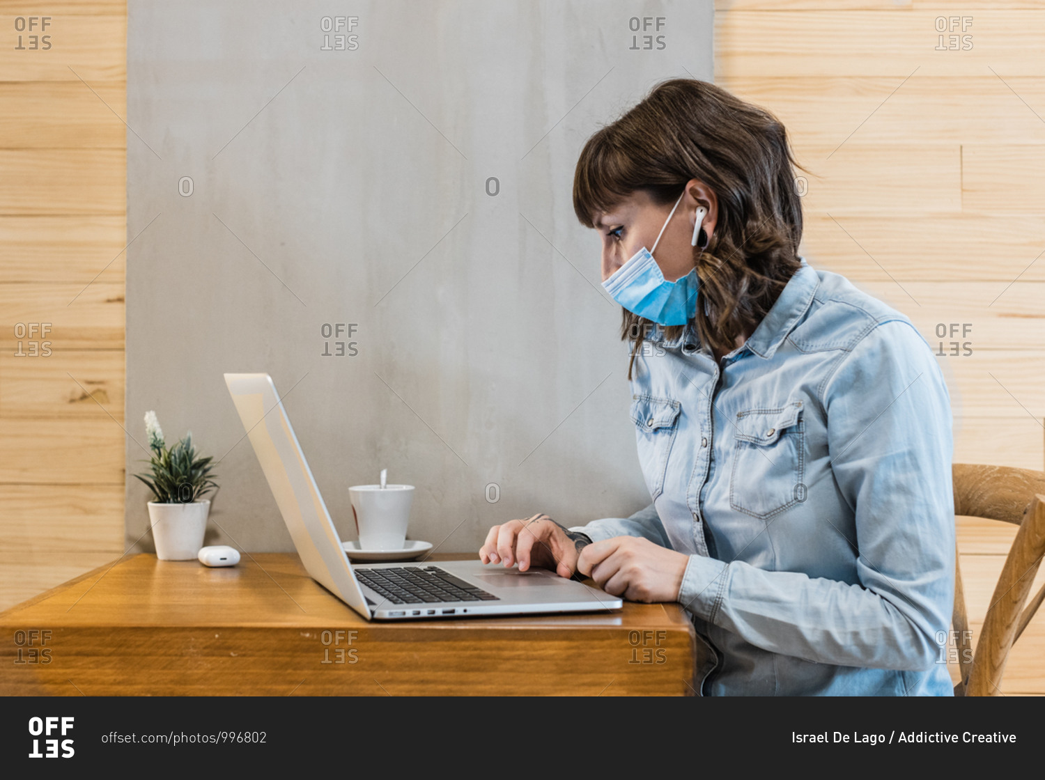 Focused female freelancer in denim shirt and medical mask using earbuds and working remotely in cafe using laptop during coronavirus epidemic