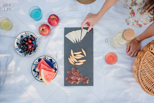 Top view of crop anonymous woman picking slice of parmesan from board with prosciutto and seeds while sitting on white cloth with fruit and lemonade during summer picnic