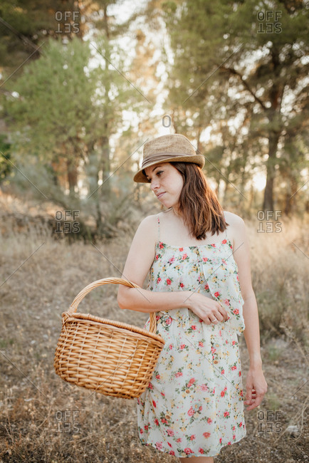 Charming woman in sundress and hat with wicker basket over arm standing on picnic blanket in back lit in summer park and looking away