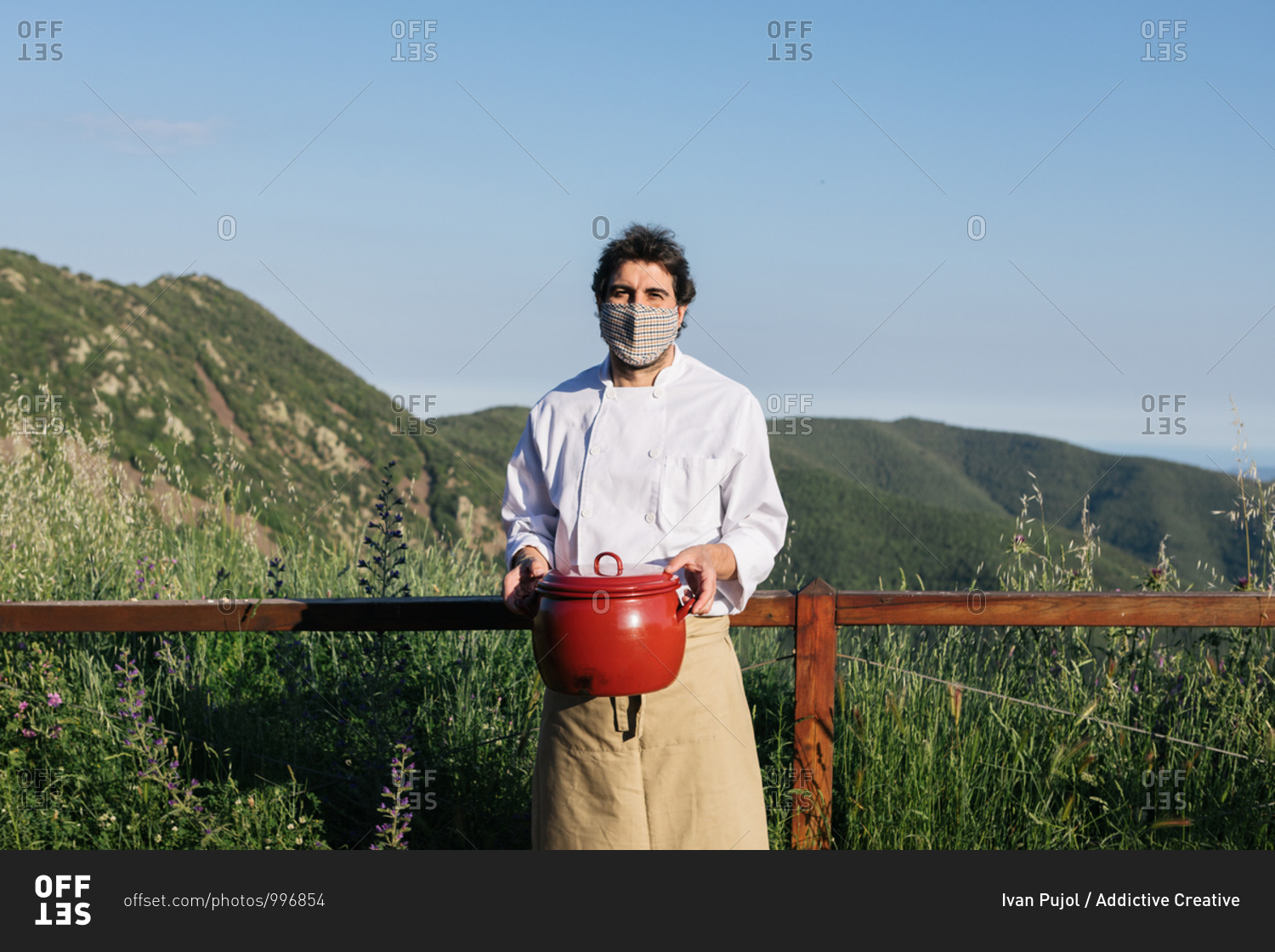 Anonymous male cook in apron medial mask standing against picturesque mountains in countryside with saucepan in hands